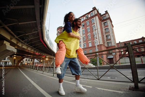 Fototapeta Multiethnic couple laughing while young woman riding piggyback on man outdoors near urban buildings, both smiling and looking forward, expressing friendship and joy