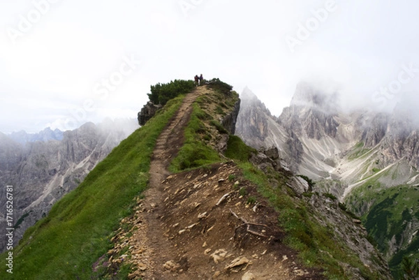 Fototapeta Hikers on a rugged mountain trail surrounded by breathtaking peaks and misty clouds in the Dolomites