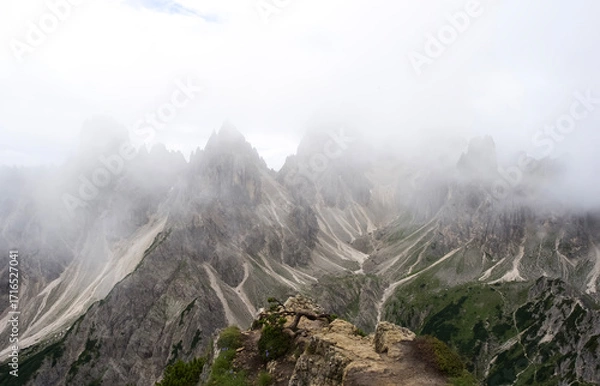 Fototapeta Majestic mountain peaks shrouded in mist with rocky terrain and lush greenery in the foreground