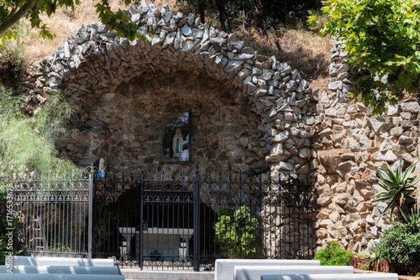 Fototapeta Stone church arch with a statue inside, surrounded by greenery. The structure is enclosed by black wrought iron gates and is shown on a bright sunny day.
