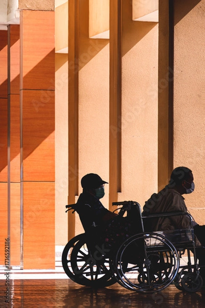 Fototapeta Silhouette 2 mature men with disability on wheelchairs are waiting for taxi in the waiting service area outside the hospital building to go home.