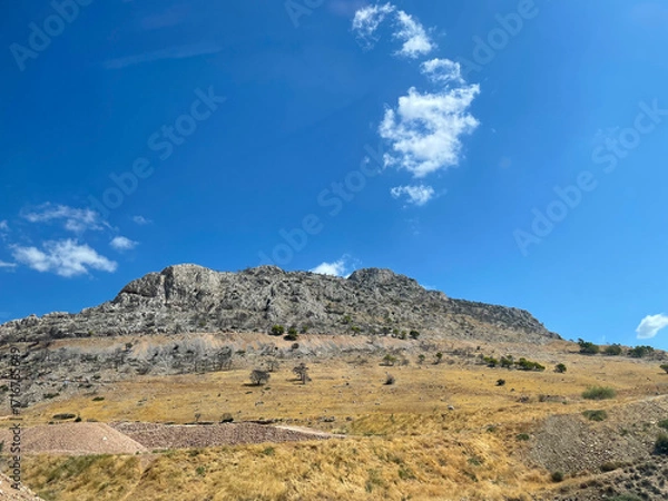 Fototapeta Rocky mountain slope under a bright blue sky with scattered clouds in Greece. Dry yellow grass and rugged stone landscape on a sunny summer day. Natural Mediterranean scenery and geology view.
