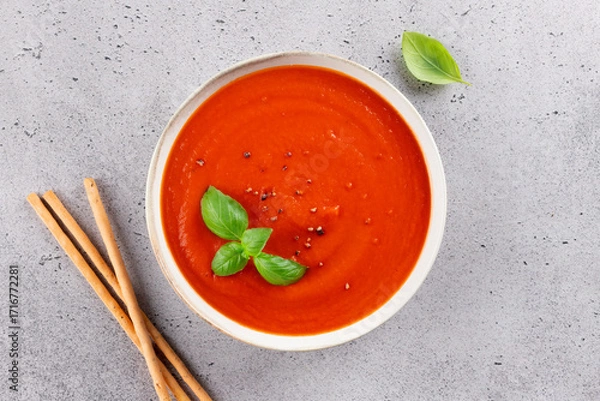 Fototapeta Tomato cream soup with fresh basil and breadsticks on a light stone background. Top view. Healthy vegetarian meal.