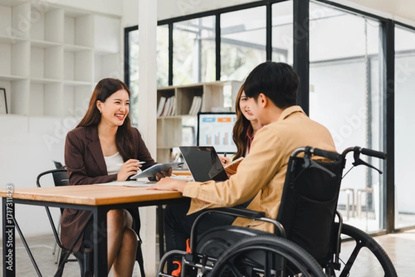Fototapeta Young man in wheelchair collaborating with two colleagues at table disability teamwork meeting laptop notebook conversation focused professional