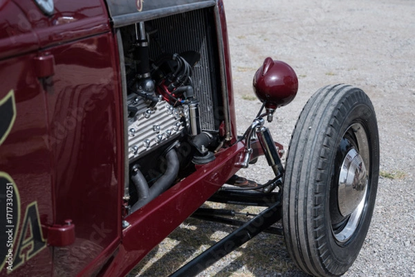 Fototapeta A close-up of the exposed V8 engine and front wheel of a classic American hot rod. The detailed shot highlights the mechanical parts and chrome suspension