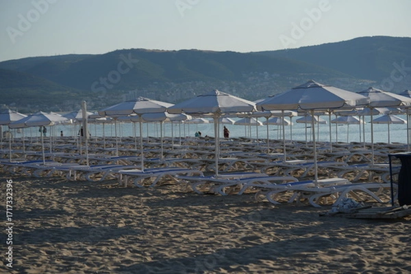 Fototapeta Beach with rows of white umbrellas and sunbeds by the sea – summer holiday resort background with mountains view, tourism and travel concept, seaside vacation destination and coastal leisure landscape