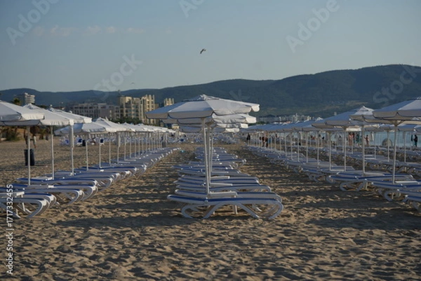 Fototapeta Beach with rows of white umbrellas and sunbeds by the sea – summer holiday resort background with mountains view, tourism and travel concept, seaside vacation destination and coastal leisure landscape