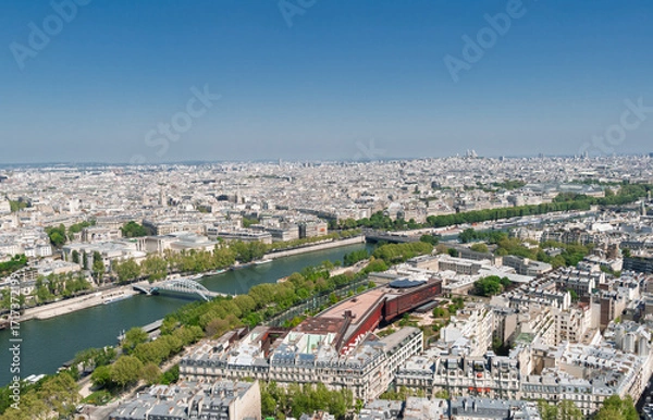 Fototapeta Paris cityscape showcasing the Seine river winding through historical buildings, green embankments, and the distinct architecture of the Quai Branly Museum under a clear sky