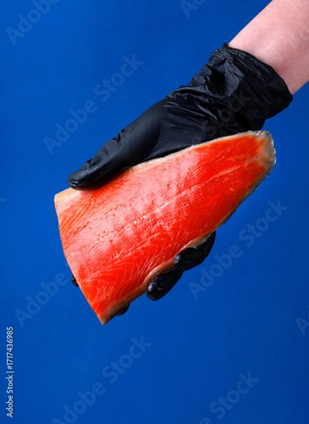 Fototapeta Cold smoked trout fillet held in hand wearing a black glove on blue background. Copy space