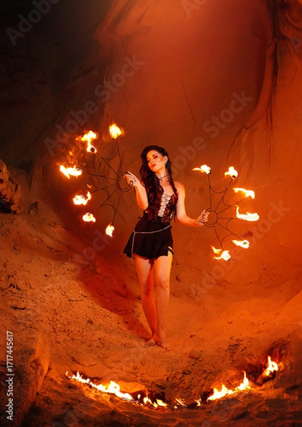 Fototapeta A young woman dressed in leather clothes stands on the sand and a flame of fire next to her. Fire show in the desert.