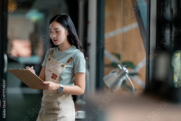Fototapeta Asian woman small business owner or employee working in cafe, taking orders, managing tasks with clipboard