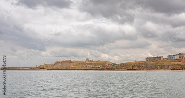 Fototapeta A seaside panorama with the ruins of an abbey on a hill and a lighthouse on a pier.