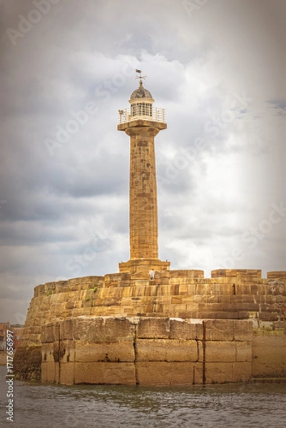 Fototapeta A lighthouse of stone material is at the end of a pier with a cloudy sky above.