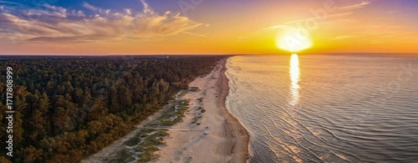 Fototapeta A breathtaking aerial view of a serene beach at sunset, where the golden sun reflects on calm waters and vibrant hues paint the sky over dense forested land