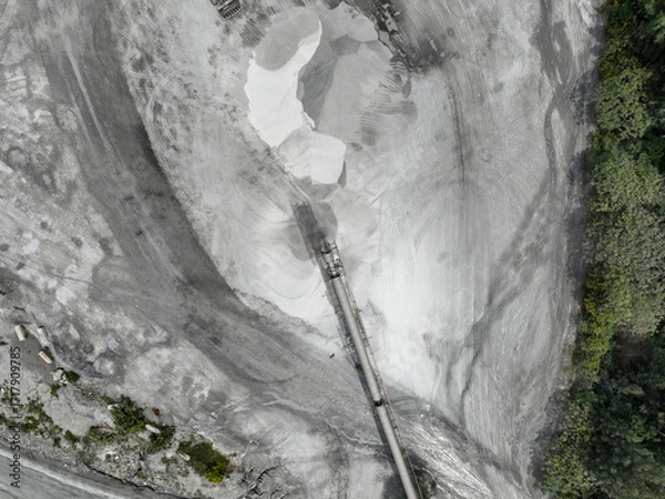 Fototapeta Industrial quarry crusher facility from above with conveyor belts and raw material piles, surrounded by erosion patterns and forest edge, symbolizing resource extraction and environmental pressure.