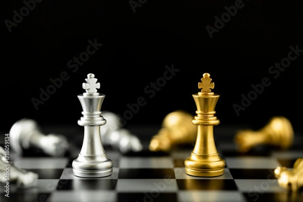 Fototapeta Golden and silver chess king pieces facing each other on a chessboard with fallen pieces, dark background.
