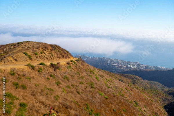 Fototapeta A wide-angle, high-angle view captures the scenic Pacific Palisades coastline from a dry hillside trail, with a low-lying marine layer of fog rolling over the Pacific Ocean below a clear blue sky in S