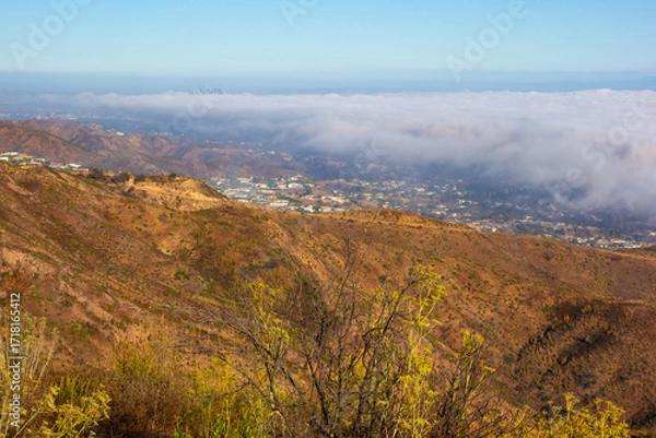Fototapeta A wide-angle, high-angle view captures the scenic Pacific Palisades coastline from a dry hillside trail, with a low-lying marine layer of fog rolling over the Pacific Ocean below a clear blue sky in S
