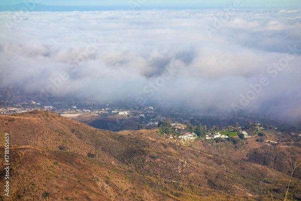 Fototapeta A wide-angle, high-angle view captures the scenic Pacific Palisades coastline from a dry hillside trail, with a low-lying marine layer of fog rolling over the Pacific Ocean below a clear blue sky in S