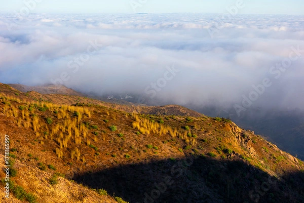 Fototapeta A wide-angle, high-angle view captures the scenic Pacific Palisades coastline from a dry hillside trail, with a low-lying marine layer of fog rolling over the Pacific Ocean below a clear blue sky in S