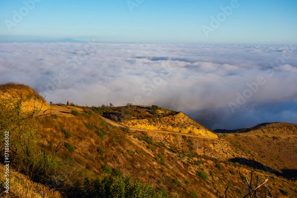Fototapeta A wide-angle, high-angle view captures the scenic Pacific Palisades coastline from a dry hillside trail, with a low-lying marine layer of fog rolling over the Pacific Ocean below a clear blue sky in S
