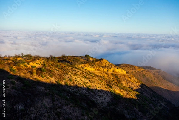 Fototapeta A wide-angle, high-angle view captures the scenic Pacific Palisades coastline from a dry hillside trail, with a low-lying marine layer of fog rolling over the Pacific Ocean below a clear blue sky in S
