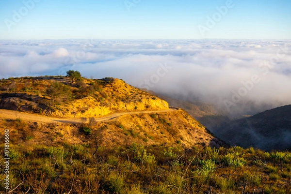 Fototapeta A wide-angle, high-angle view captures the scenic Pacific Palisades coastline from a dry hillside trail, with a low-lying marine layer of fog rolling over the Pacific Ocean below a clear blue sky in S