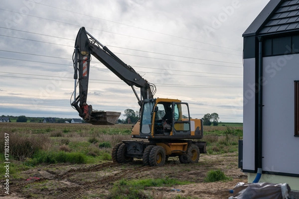 Fototapeta Wheeled Excavator at Rural Construction Site with Bucket Raised and Open Cabin.
