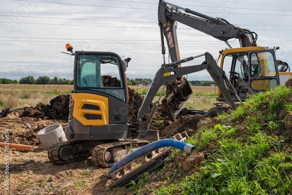 Fototapeta Two excavators at a construction site digging soil for underground pipe installation in a rural area.