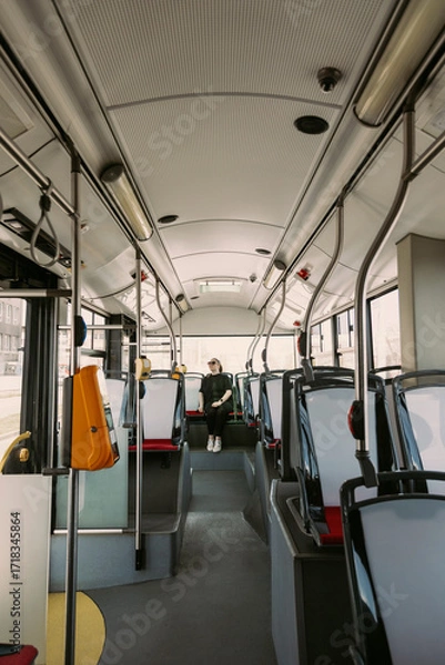 Fototapeta Wide-angle view of a stylish woman in sunglasses sitting alone at the back of a bright, empty city bus. Great for depicting urban commuting, independence, and quiet transport moments.
