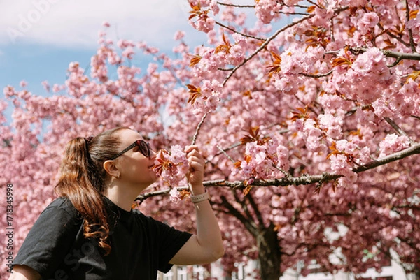 Fototapeta A smiling woman with a ponytail smells blooming cherry blossoms in a sunny park. Perfect for lifestyle content, spring ads, wellness themes, floral branding, and nature-inspired visuals.