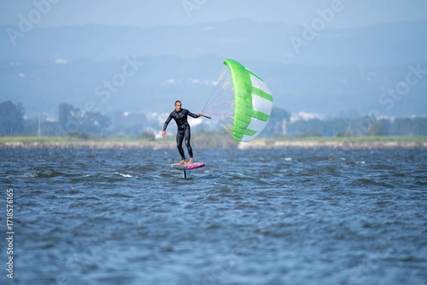 Fototapeta A middle-aged man in a wetsuit glides on a hydrofoil board with a green and white kite