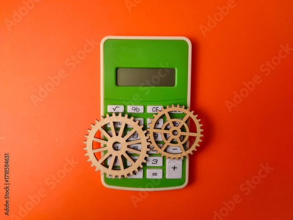 Fototapeta The image displays a green calculator accompanied by two wooden gears on an orange background