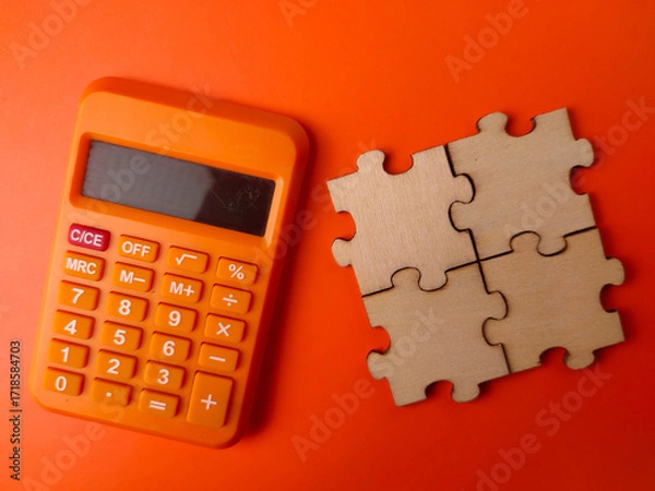 Fototapeta The image displays a small orange electronic calculator alongside four interlocking wooden puzzle pieces on an orange background