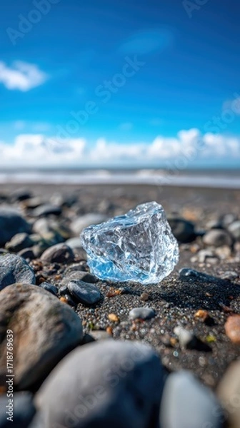 Fototapeta A glistening chunk of ice rests on a rocky beach with soft waves and a clear blue sky dotted with clouds in the background landscape scene.