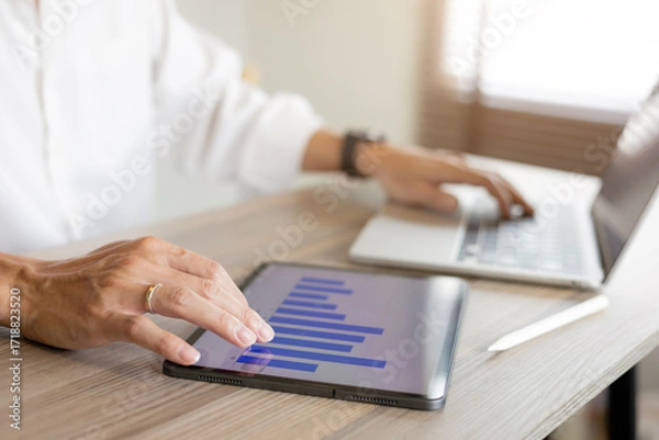 Fototapeta Businessman analyse on financial data using tablet with bar chart and laptop on wooden desk. Concept of digital marketing, business growth, investment strategy and data analytics