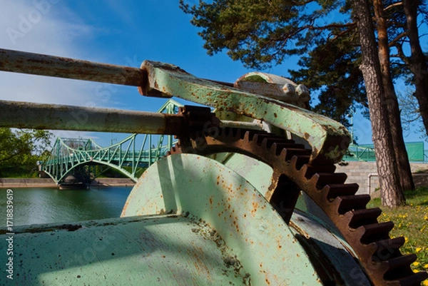 Fototapeta Close-up of an old rusty gear mechanism of a drawbridge with a metal bridge in the background. Industrial engineering detail.