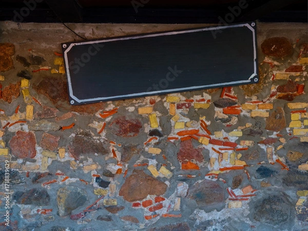 Fototapeta Empty black signboard on an old stone brick wall with rough texture, ready for text or logo placement.
