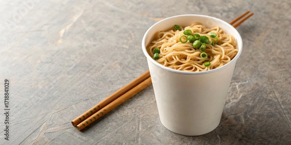 Fototapeta Instant noodle cup with chopstick and green onion garnish on textured surface, warm and appetizing meal