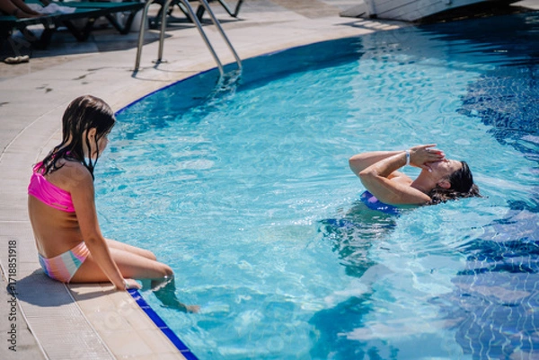 Fototapeta Woman enjoying a swim while a young girl sits at the edge of a clear blue swimming pool during a sunny summer day at a resort.