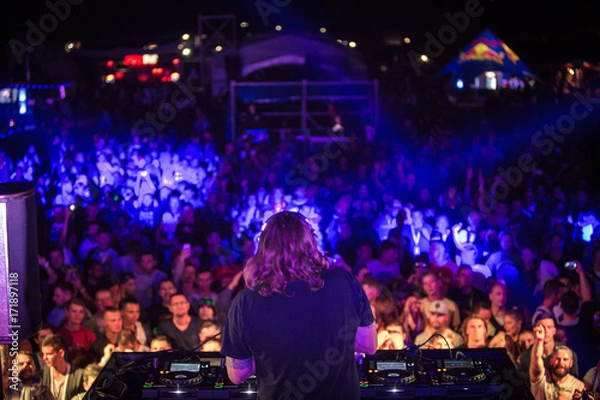 Fototapeta DJ with headphones at night club party under the colorful light and people crowd in background. Dj at open aire party. Night party at sea coast. Beach party.