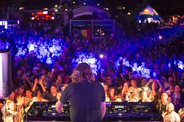 Obraz DJ with headphones at night club party under the colorful light and people crowd in background. Dj at open aire party. Night party at sea coast. Beach party.