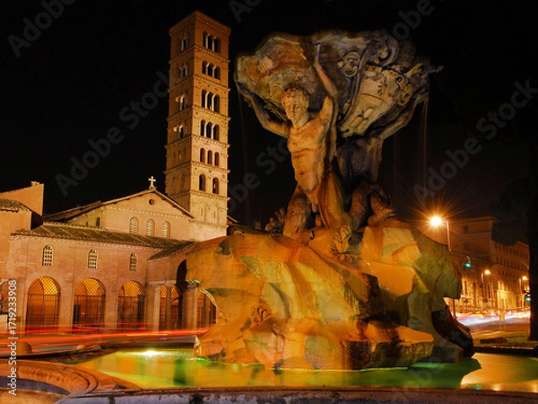 Fototapeta Beautiful baroque Fountain of the Tritons with medieval Basilica of Saint Mary in Cosmedin lit up at night in Rome