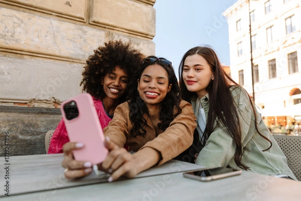 Fototapeta A group of three female friends are sitting at a table and smiling while looking at a phone that one of them is holding