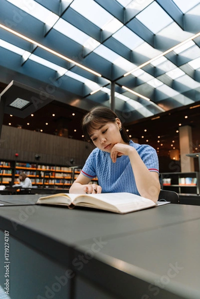 Fototapeta A female student is reading a book and holding a pen while resting her head on her hand and sitting at a table