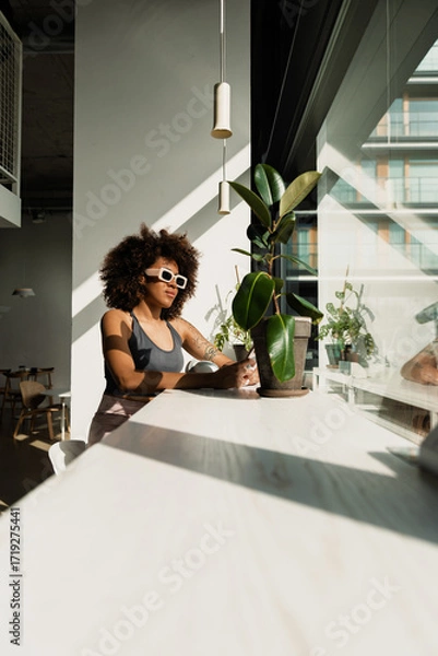 Fototapeta A woman is sitting at the table and looking out the window while holding the phone