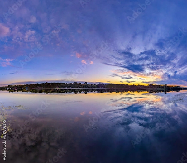 Fototapeta Sunset over calm river shore panoramic view. Dramatic sunset violet dark. Yellow Orange blue color shades cloudy sky. Clouds reflected quiet water. A strip of forest silhouette fills horizon line.