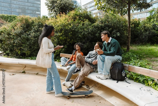 Fototapeta A female student stands and holds a phone while listening to a group of three students sitting across from her and smiling