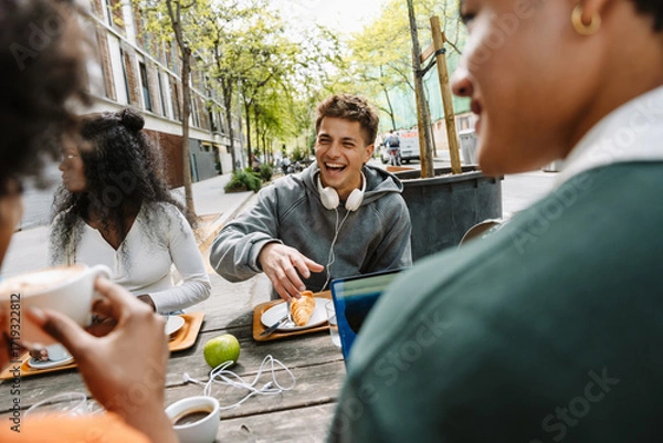 Fototapeta A male student laughs while sitting at a table with a group of three students during lunch