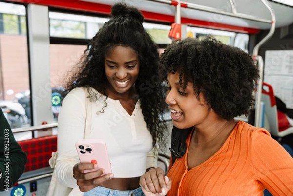 Fototapeta Two female students stand on public transport and laugh while looking at the phone one of them is holding